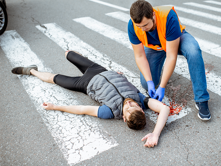 A young white male in jogging clothes lays in a crosswalk while another young white male in a safety vest pretends to attend to bleeding wounds. 