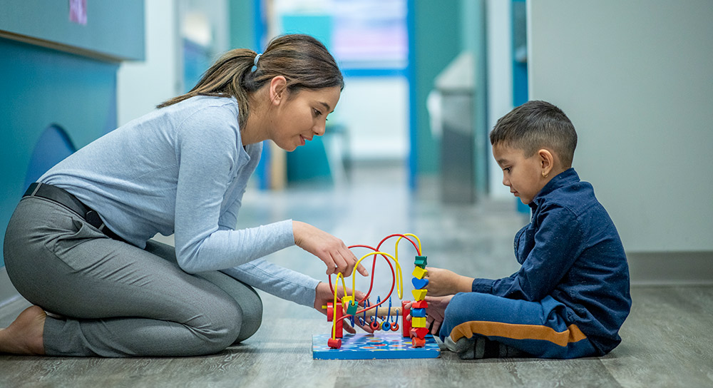 1000x544_Young-teacher-plays-on-the-floor-with-small-boy A young Latina sits on the floor opposite a Latino toddler. In between them is a colorful toy that they both are invested in.