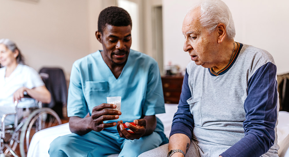 Two men sit on a hospital bed: a younger, Black healthcare worker in scrubs hands medication to an older white man.