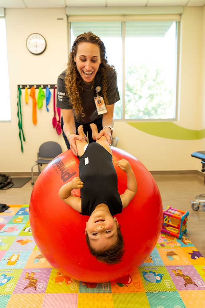 full_size-PediatricRehab-3 A healthcare worker holds a toddler boy by his legs as he lays back on a giant, inflatable ball. They are in a colorful, child-friendly rehabilitation room.