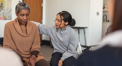 Neuroimmunology-Support a young Black woman consoles an older Black woman who is sitting in a chair clasping her hands together