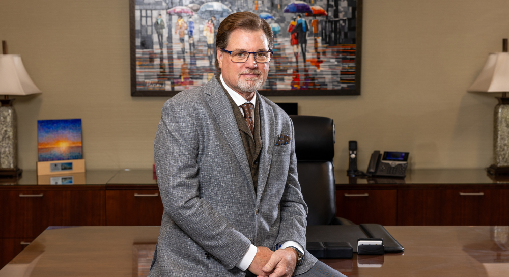 A white man with brown hair, wearing a gray suit, sits on the edge of a desk in a nice office. 