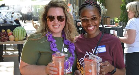 Two young women hold Community-labeled cups with cold beverages outside at a farmers market