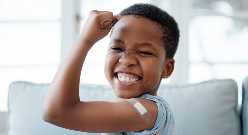 Un niño pequeño, de unos 7 años, flexiona el brazo y sonríe a la cámara. Tiene un apósito en el bíceps.