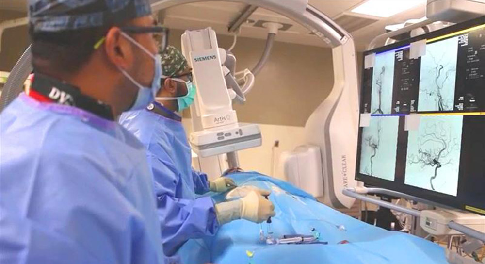 Two male doctors in scrubs work over a patient. Monitors show scans of a brain.