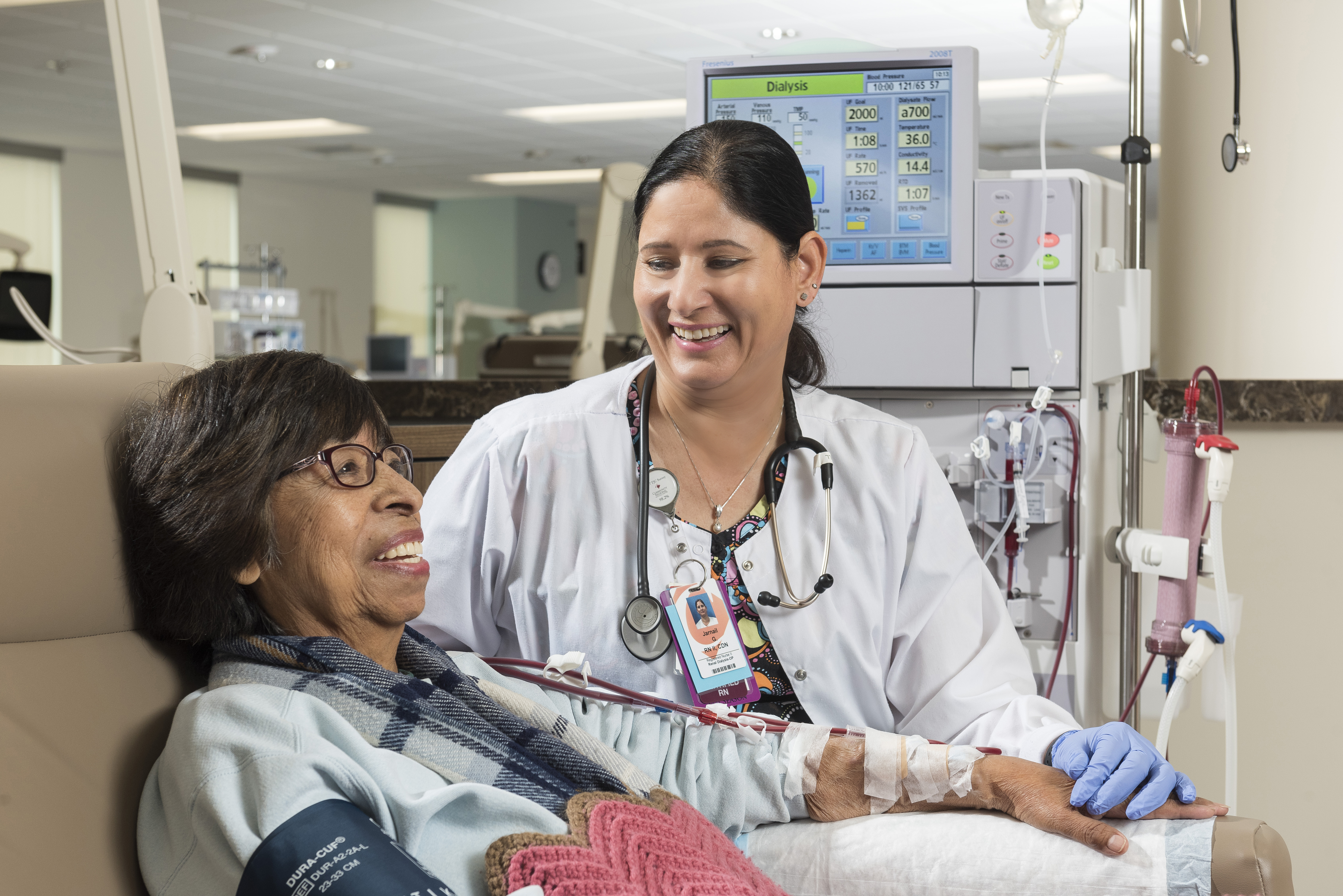 An older woman hooked up to a dialysis machine smiles as a technician holds her hand