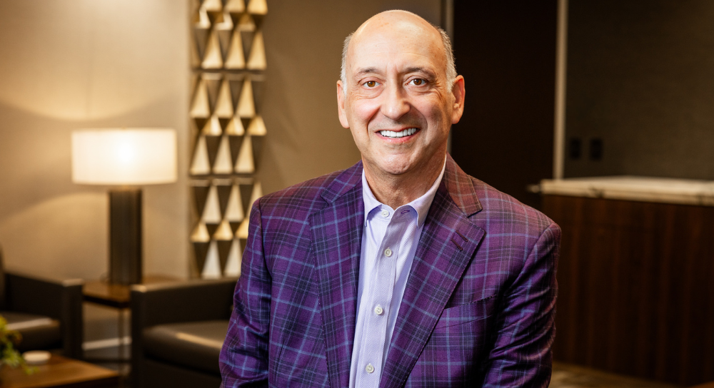 A man in a purple blazer sits in a comfortably lit office. zHe is smiling at the camera.