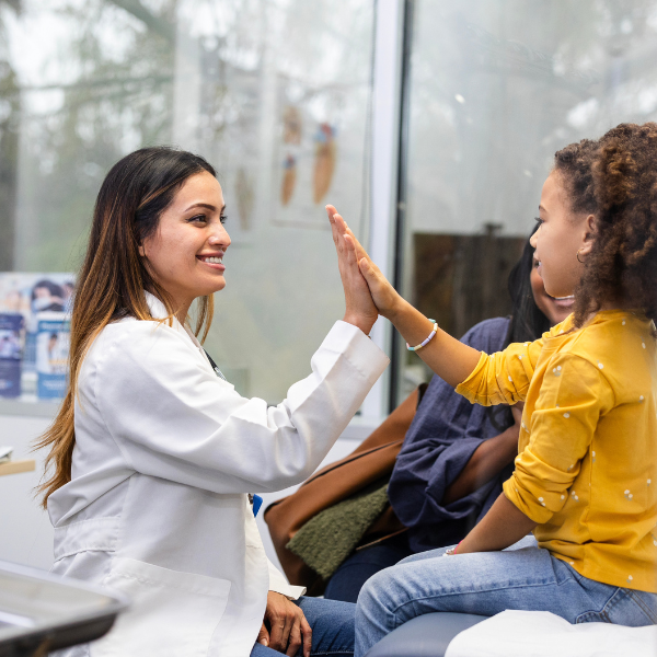 A young Latina doctor high fives her patient, a Black tween girl in a yellow shirt