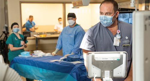 Five people in surgical scrubs are spread around a hybrid OR