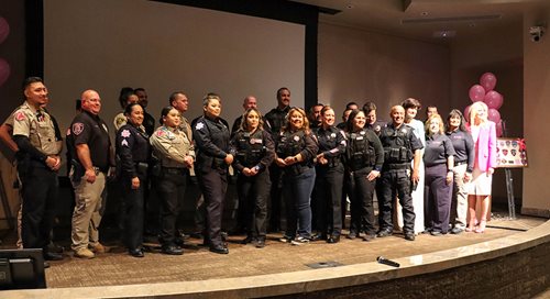 A group of about 25 officers with law enforcement stand on a stage, each wearing their respective pink patches on their uniforms.