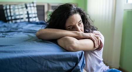 A woman with long curly brown hair sits on the floor and leans onto her bed, a distressed look on her face