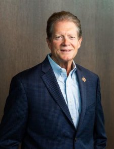 An older white man looks at the camera. He is wearing a blue blazer and blue shirt and is standing in front of a dark wood-paneled wall.