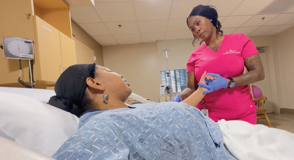 A female doula in pink scrubs holds the hand of a pregnant woman in a hospital bed