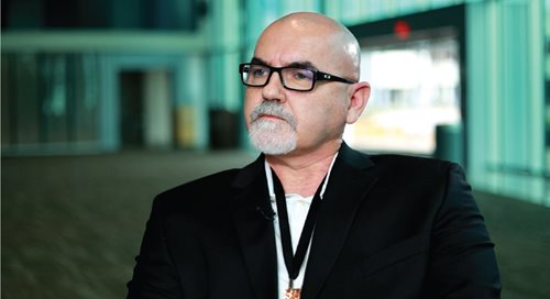 Tom Minas, wearing black-framed glasses and a dark blazer, is shown from the chest up, sitting in a hospital lobby