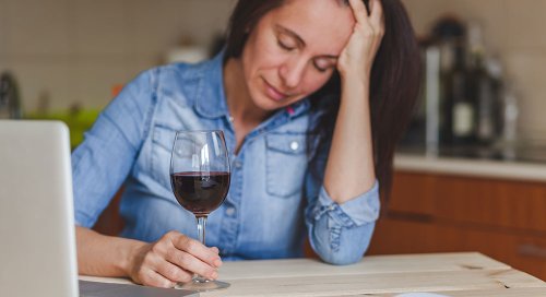 A white woman in her 30s with long brown hair holds her head in her hand. Before her is a glass of red wine.